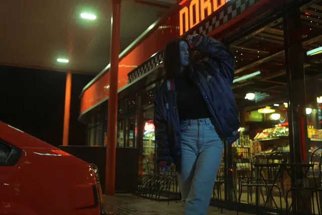 A woman standing outside a diner with neon lights at night, wearing a jacket and jeans.