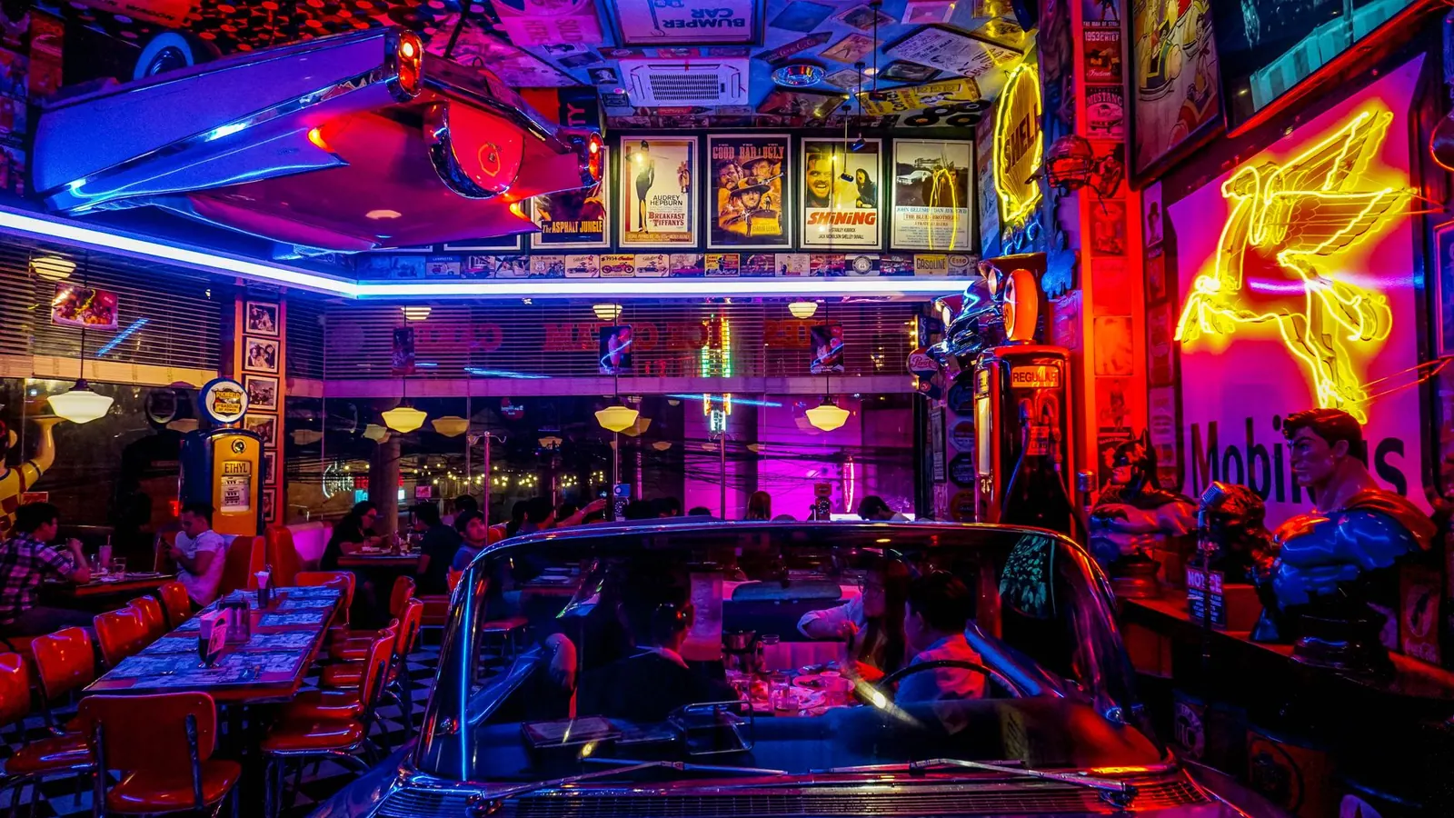 A lively retro diner bustling with people, neon lights, and classic car decor, set at night.