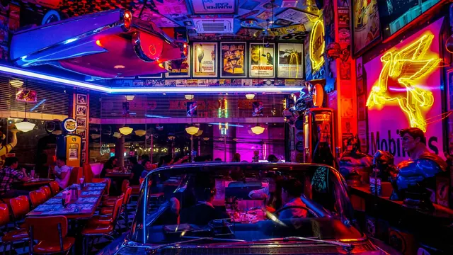 A lively retro diner bustling with people, neon lights, and classic car decor, set at night.