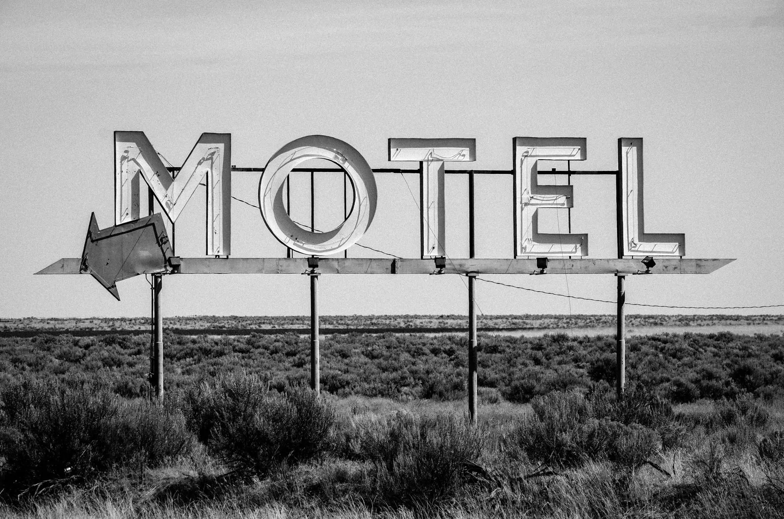 Black and white image of an old motel sign with an arrow in a grassland setting.